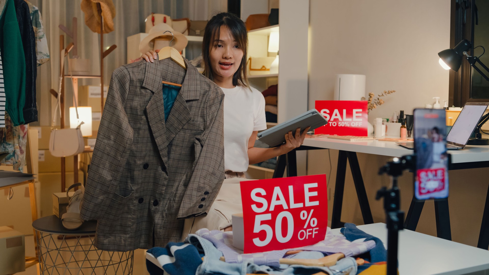 Young Asian woman live streaming on phone show new collection fashion clothes to customer in shop at night. SME small business e-commerce.